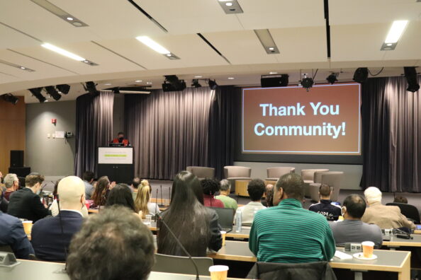 A group of people sit in an auditorium while listening to a speaker. On the stage is an orange and white sign that states, "Thank you Community!"