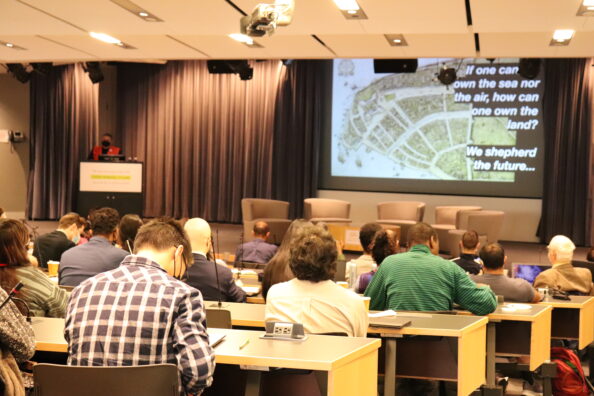 Audience members sit in an auditorium. On the mainstage is a projection screen that says: "If one can own the sea nor the air, how can one own the land? We shepherd the future..."