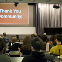 A group of people sit in an auditorium facing an orange slide that reads "Thank You Community!" in white text.