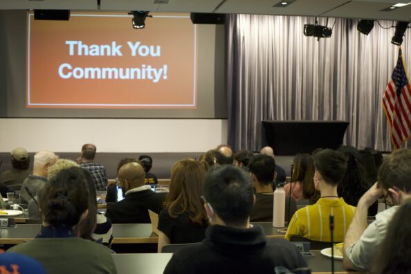 A group of people sit in an auditorium facing an orange slide that reads "Thank You Community!" in white text.